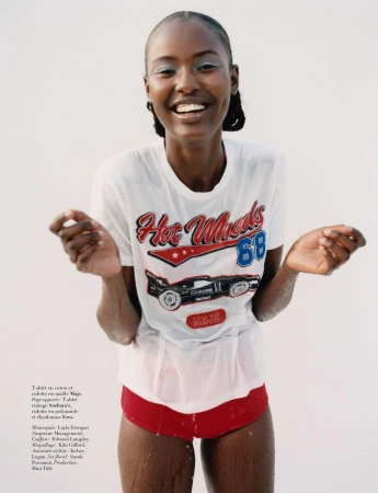 A smiling African-American woman in a wet t-shirt with a racecar graphic and red shorts poses in front of a white background.