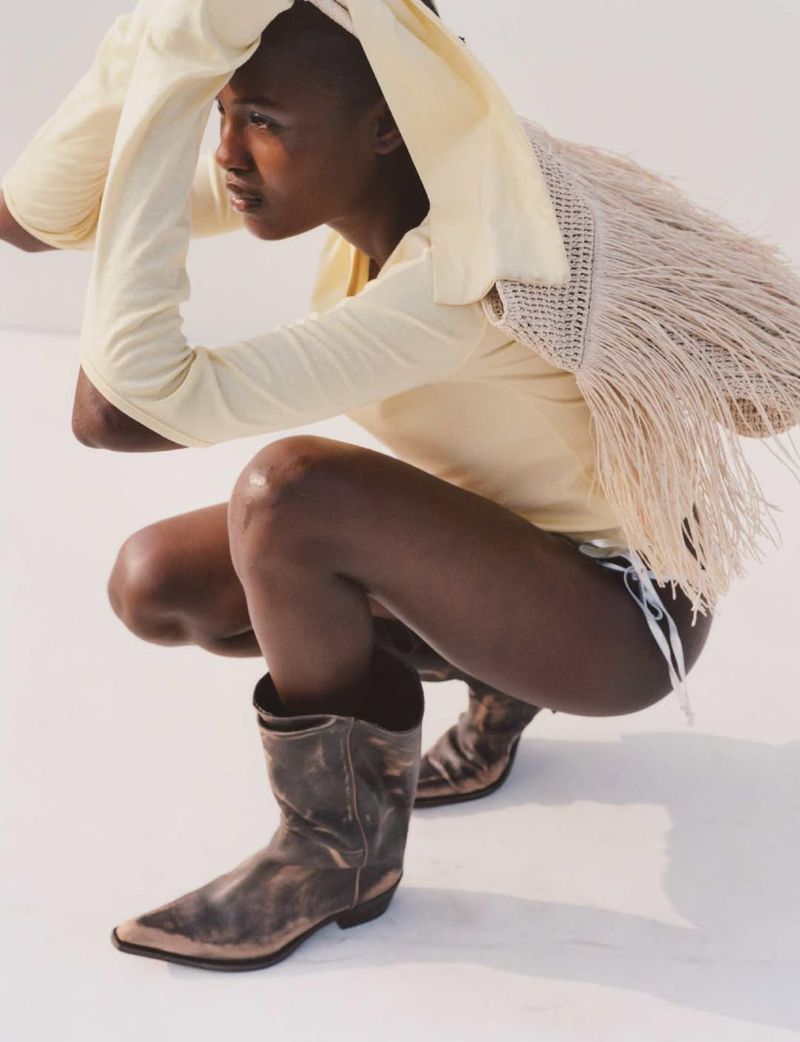 Young adult in a light-yellow top and cowboy boots crouching indoors on a white surface in studio lighting, looking away from camera.