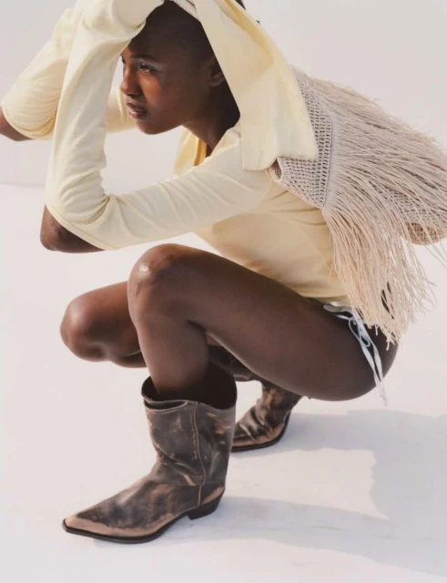 Young adult in a light-yellow top and cowboy boots crouching indoors on a white surface in studio lighting, looking away from camera.