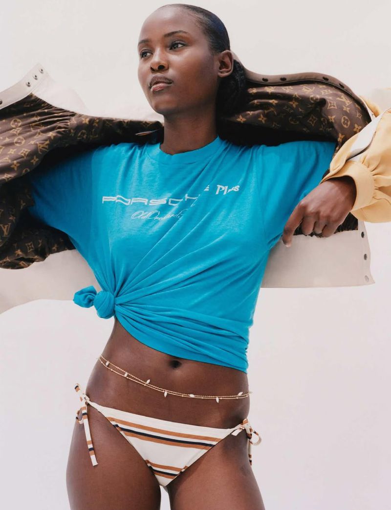Dark-skinned woman poses in a turquoise t-shirt and striped bikini bottoms, a gold chain, and jackets hanging off her shoulders in a studio setting.