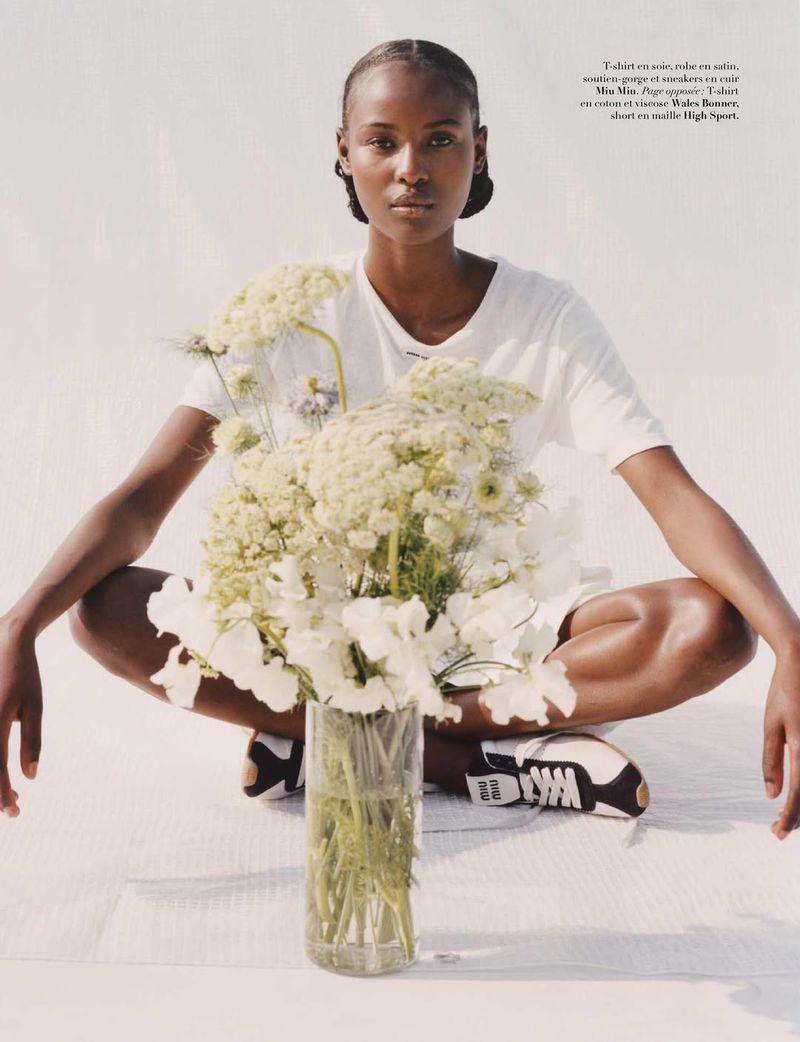 A lone, dark-skinned woman sits cross-legged in a studio with a tall bouquet in the foreground, wearing casual white t-shirt and sneakers.