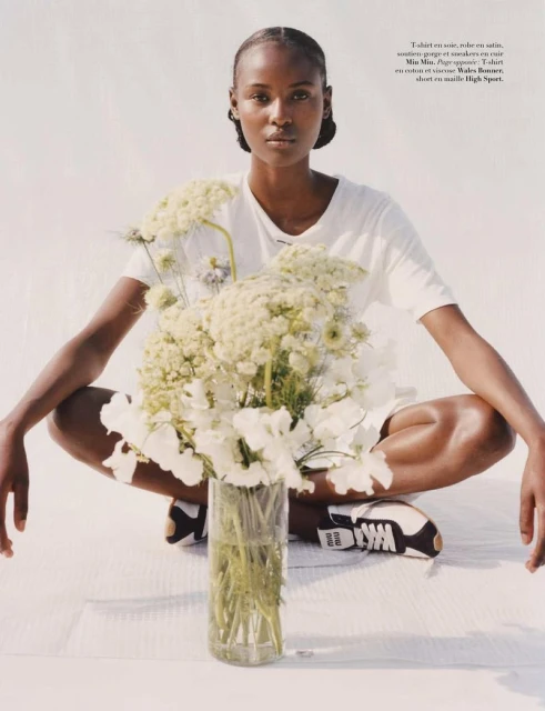 A lone, dark-skinned woman sits cross-legged in a studio with a tall bouquet in the foreground, wearing casual white t-shirt and sneakers.