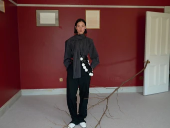 Model Lina Zhang in dark blouse and black trousers standing in a red room with a branch prop for Holiday Magazine
