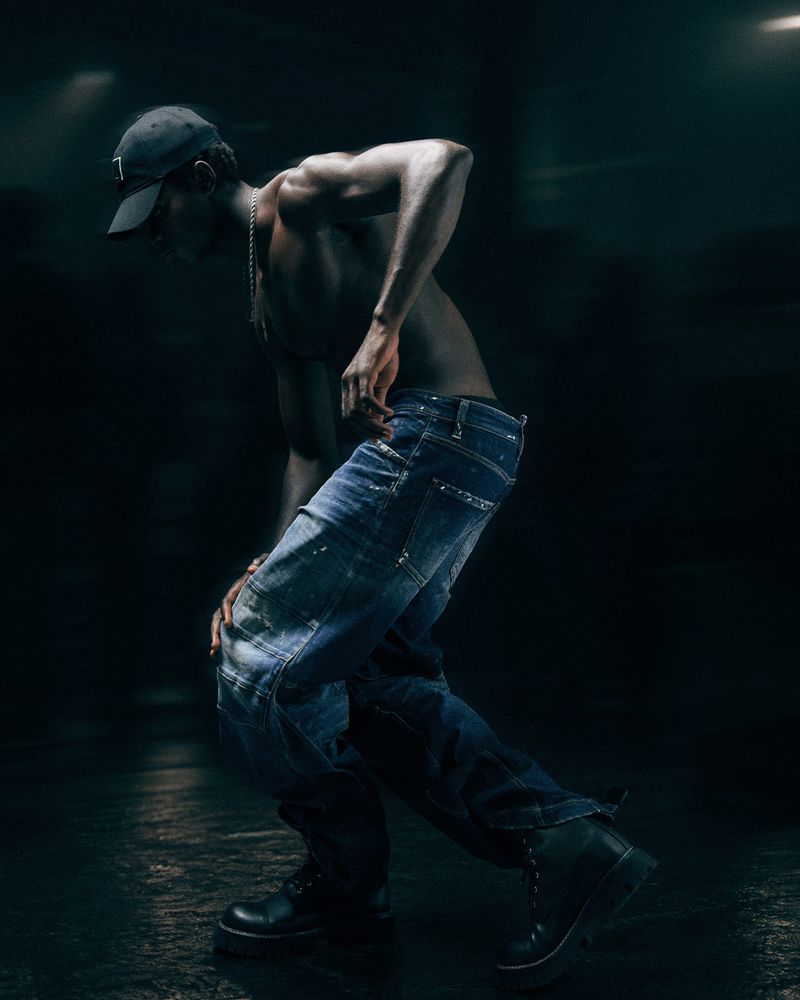 Shirtless man wearing a baseball cap and denim jeans posing in low-key lighting for GQ Turkey