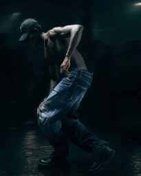 Shirtless man wearing a baseball cap and denim jeans posing in low-key lighting for GQ Turkey