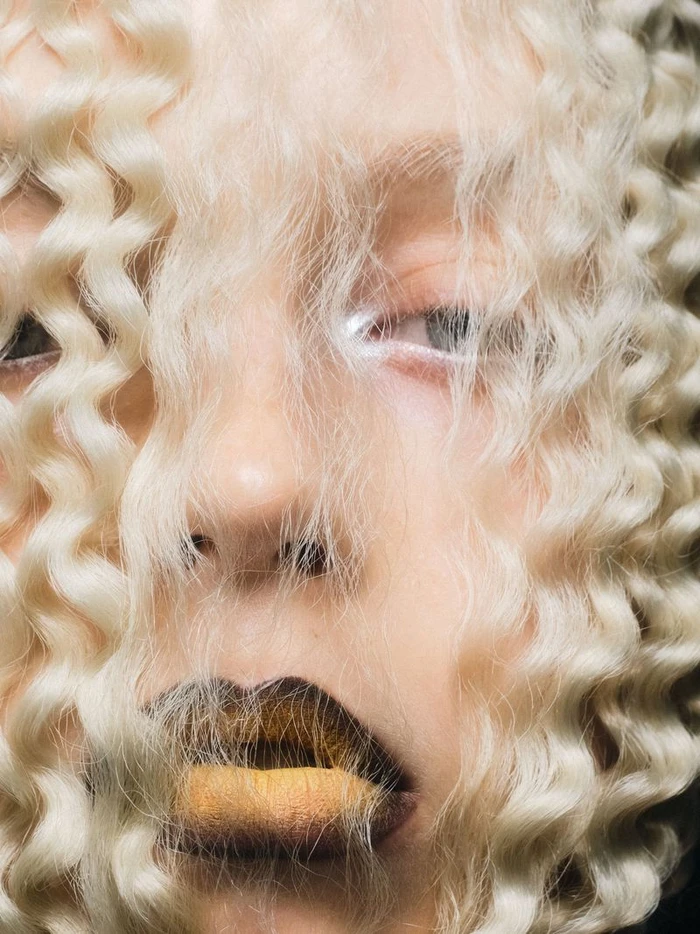 Close-up portrait of a person with tight blonde curly hair framing the face, featuring white eyeshadow and dark lips.