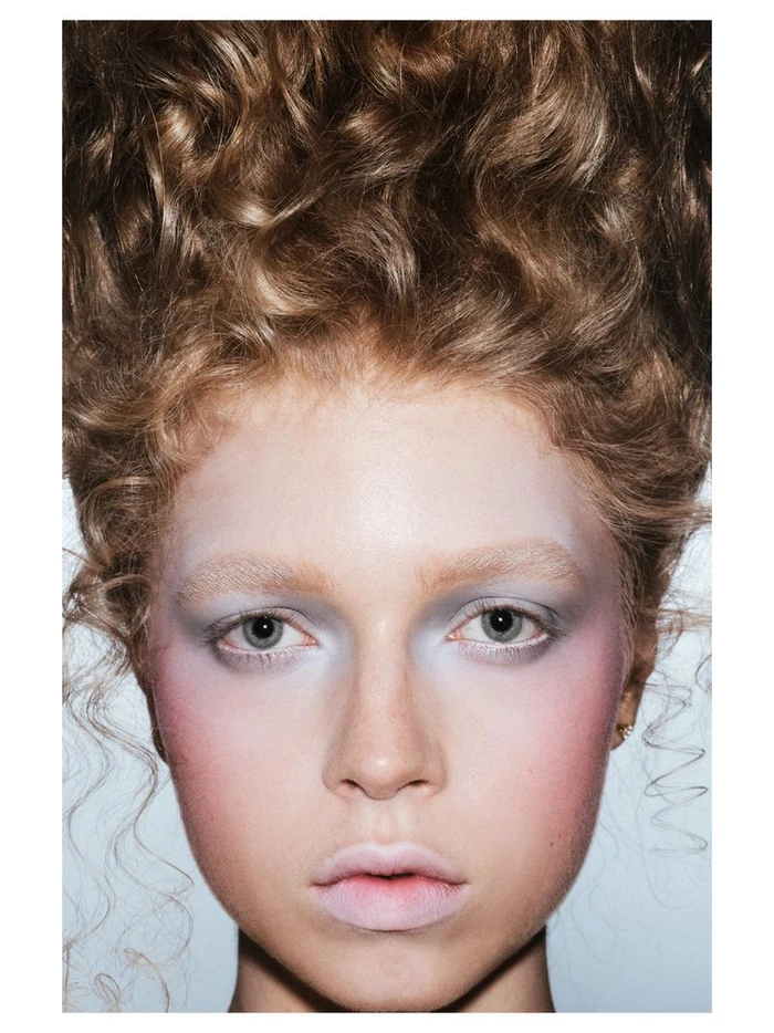 Close-up portrait of a woman with voluminous curly hair and pale pastel blue eye makeup against a white background