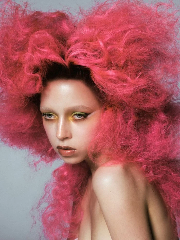 Close-up portrait of a model with voluminous bright pink hair and dramatic yellow eyeshadow against a neutral studio background.