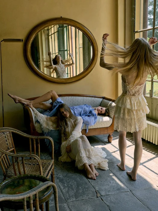 Three female models in a sunlit room, one standing and tossing her hair, two seated on a sofa and floor in an editorial shoot.