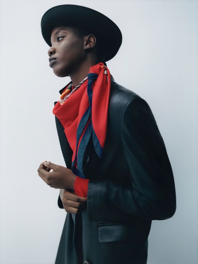 Model wearing a black hat, red scarf, and dark coat against a plain white background for Manifesto Magazine editorial