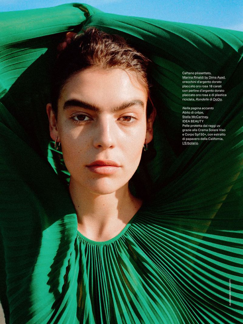 Close-up fashion shot of a model with dark hair and fair skin wearing a green pleated garment against a blue sky background.