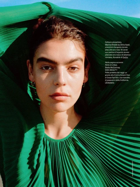 Close-up fashion shot of a model with dark hair and fair skin wearing a green pleated garment against a blue sky background.