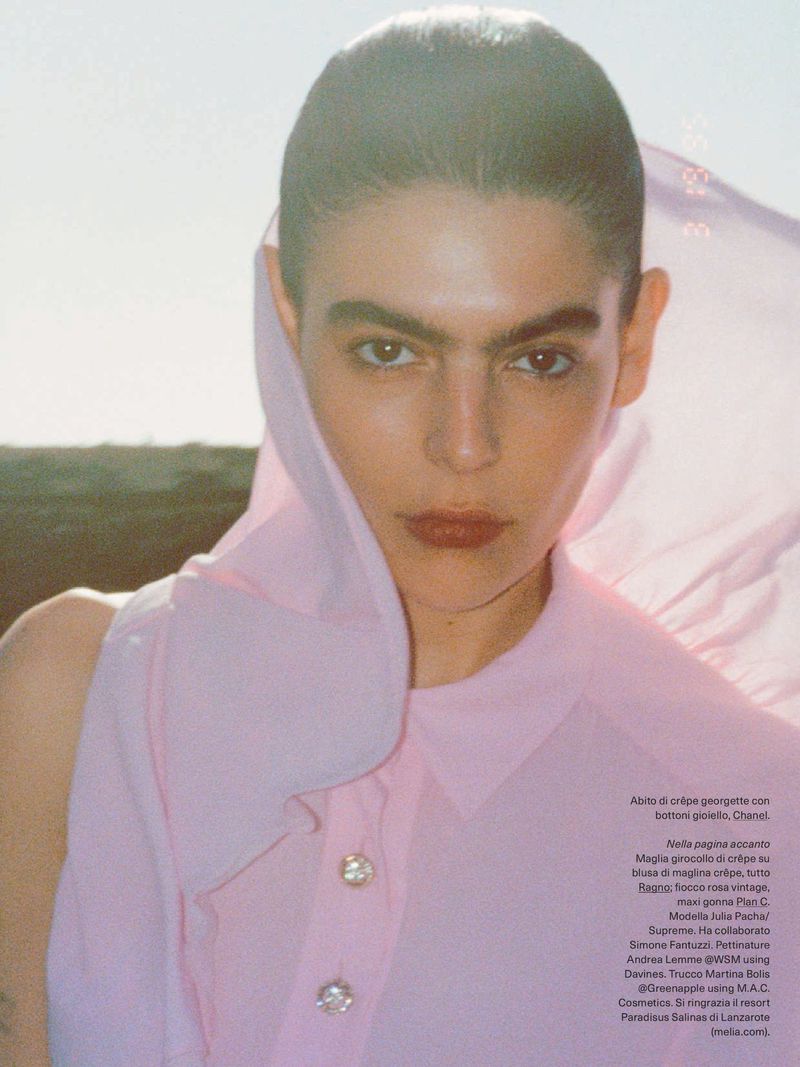 Portrait of a woman with dark hair wearing a pink blouse and hood, posing outdoors in natural light against a bright, sunny sky.