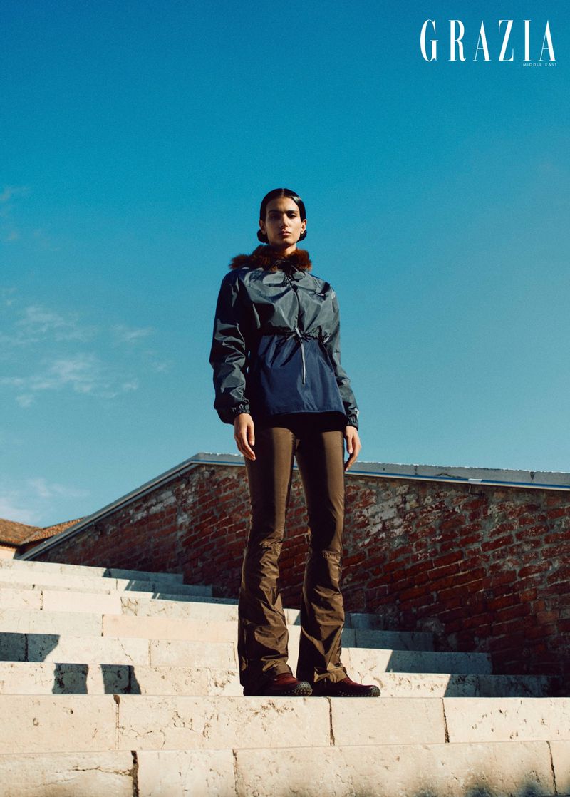 Model standing on stone steps against a clear blue sky wearing a dark hooded jacket and brown trousers for Grazia Middle East.
