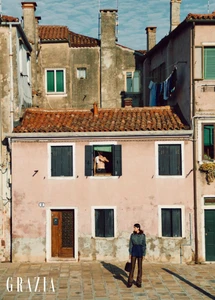 Model standing on a paved square in front of a weathered pink building with tiled roofs under an overcast sky.