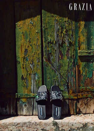 Black embellished slides placed in front of a rustic green and yellow distressed wooden door for Grazia Middle East