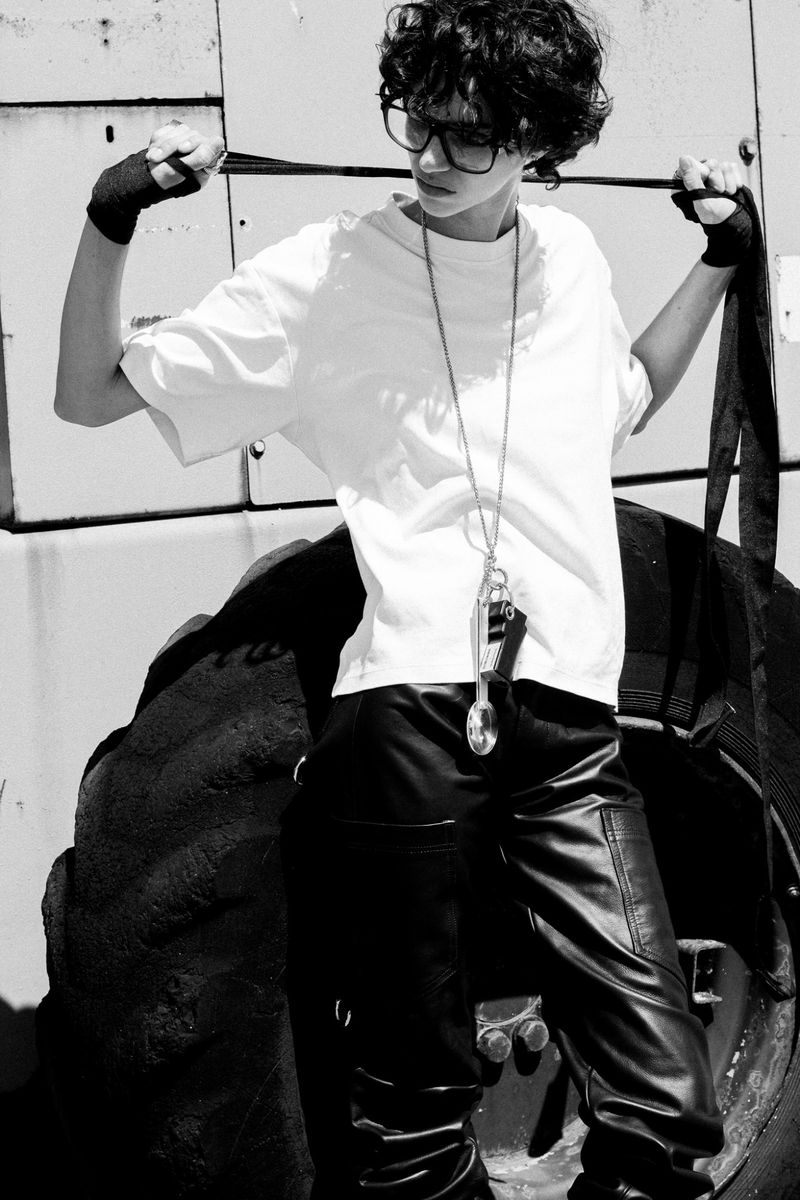 Model with dark curly hair wearing glasses, white t-shirt and black pants holding straps in front of large tires for Grey Magazine editorial.