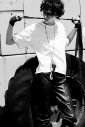 Model with dark curly hair wearing glasses, white t-shirt and black pants holding straps in front of large tires for Grey Magazine editorial.