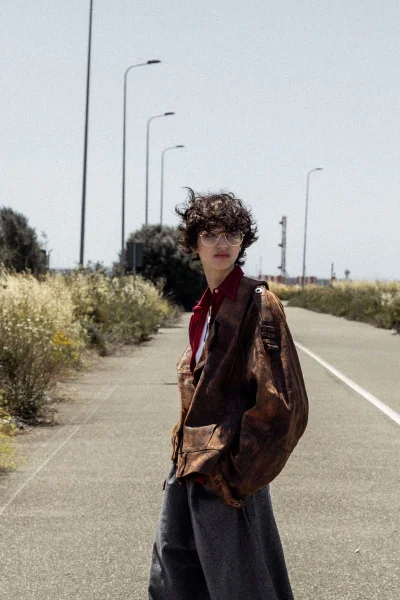 Model with dark curly hair wearing a brown jacket standing on a paved road lined with streetlights under a bright sky.