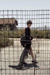 Model with short curly hair wearing black clothing behind a wire mesh fence outdoors in a desolate yard.