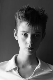 Monochrome portrait of a person with short spiked hair and septum piercing wearing a white collared shirt against a dark background