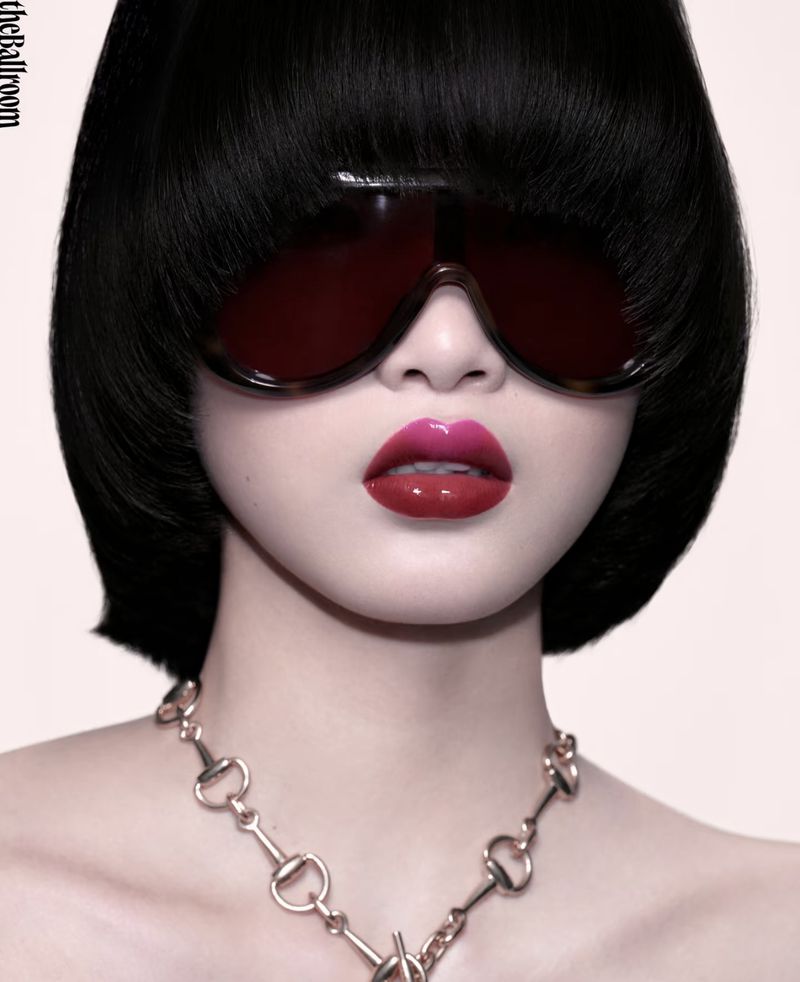 Close-up portrait of a model with short black bob hair, dark visor sunglasses, glossy red lips, and a metallic link necklace on a plain background.