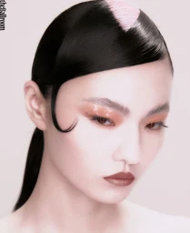 Close-up portrait of a model with sleek black hair, a pastel pink section on top, and glossy makeup against a white backdrop.