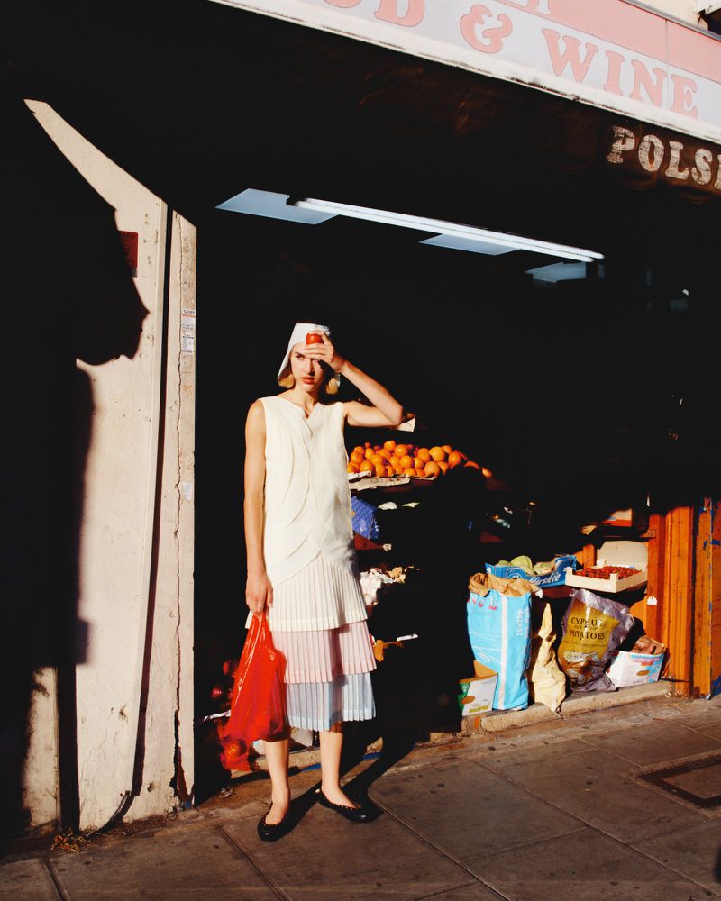 Model standing in front of a shopfront holding a red plastic bag wearing a layered skirt and white top