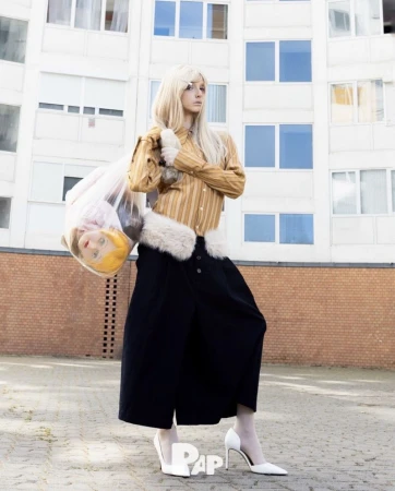 Woman in yellow striped shirt and black skirt holding a plastic trash bag with a doll head inside.