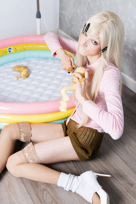 Blonde model in pink shirt and green shorts seated peeling a potato beside a small inflatable pool for PAP Magazine.