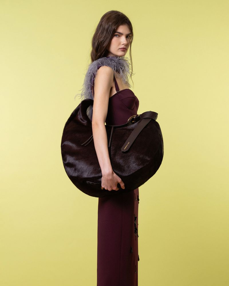 A stylish woman poses in a long plum dress, showcasing a large, dark fur purse with a shoulder strap against a pale yellow backdrop.
