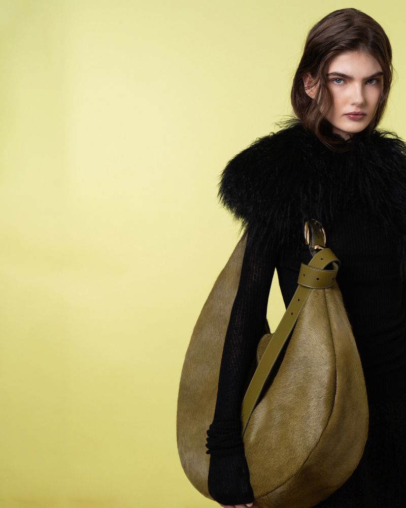 A woman poses in black clothing with a unique olive green hobo bag against a solid pastel green backdrop, creating a fashion-forward aesthetic.
