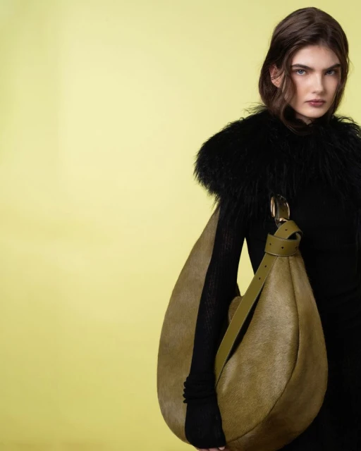 A woman poses in black clothing with a unique olive green hobo bag against a solid pastel green backdrop, creating a fashion-forward aesthetic.