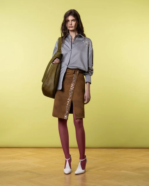 Fashionable brunette poses in a grey blouse, brown skirt with embellishments, red tights, and white heels, holding an olive green bag.