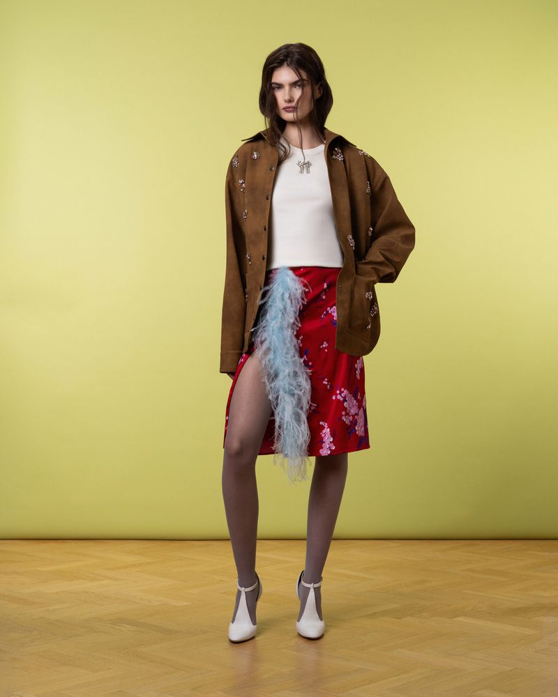 A woman in a brown jacket, white top, and red floral skirt stands against a green background with a neutral expression. She wears white T-strap heels.