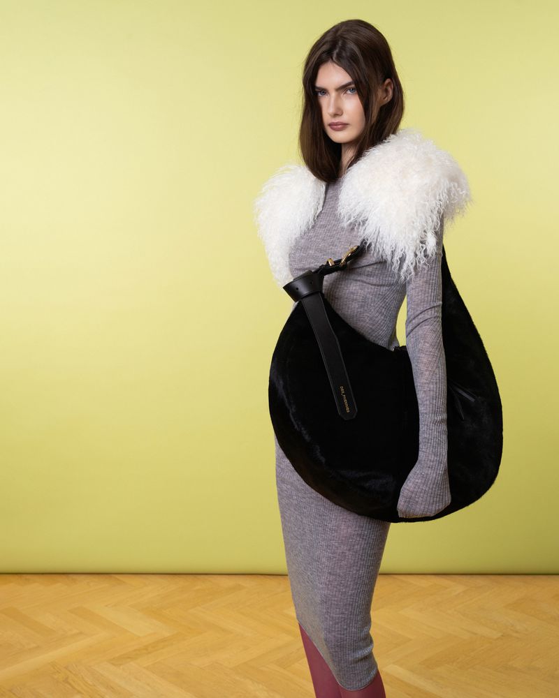 A woman poses in a studio with a grey dress, large black handbag and fur shawl against a lime green background with wood flooring.