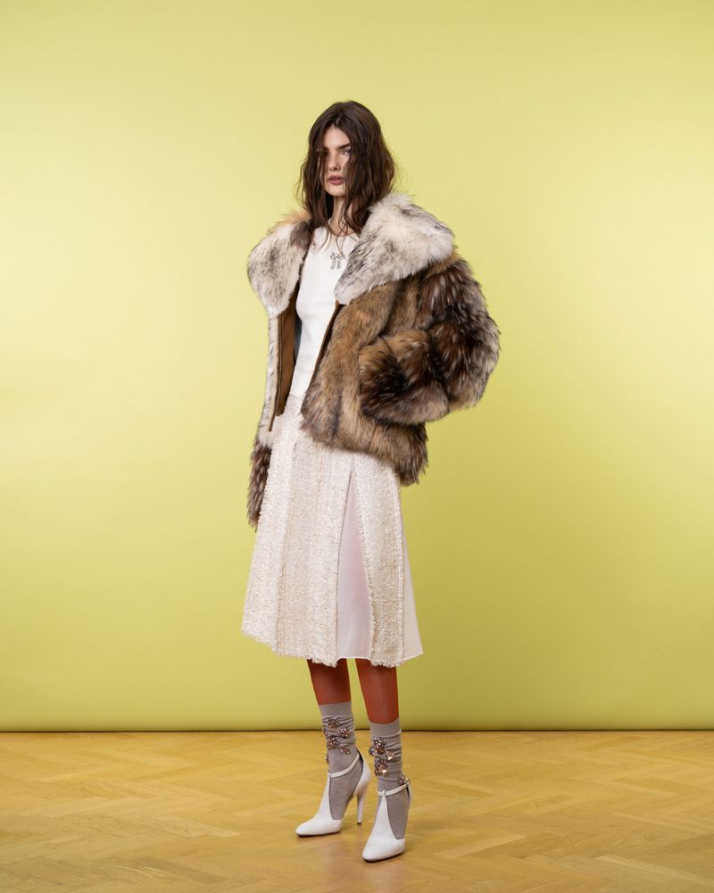 A woman stands in a photo studio wearing a fur coat, white skirt, socks with embellishments, and white heels, against a yellow background.