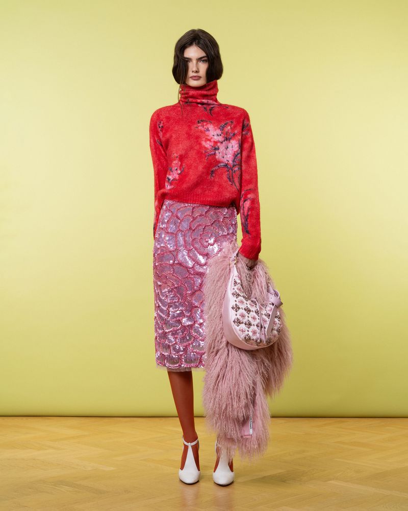 Full-length studio shot of a young adult woman posing in a red floral turtleneck sweater, pink sequin skirt, and white shoes with a pink purse.