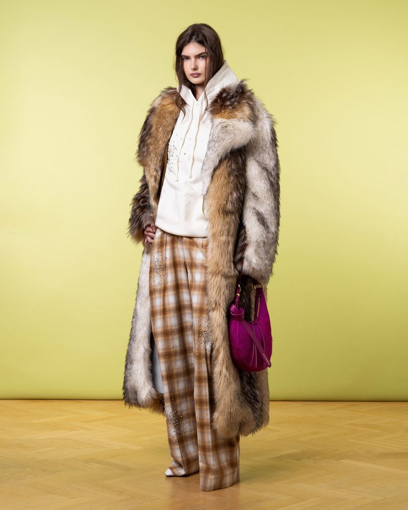 A woman in a fur coat stands in front of a yellow background, wearing plaid pants and a hoodie, complemented by a magenta purse.