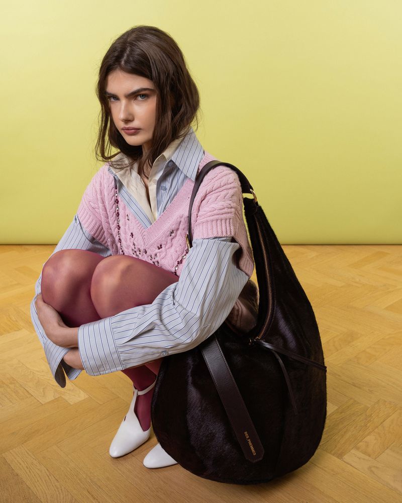 Brunette woman poses with a large dark fur shoulder bag in a studio with a yellow background and light wood floor.