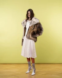 Model standing in a brown fur jacket, cream pleated skirt, embellished socks, and white heels against a yellow backdrop.