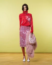 Model in red turtleneck and sequined pink skirt holding a pink feathered bag against a gradient yellow background.