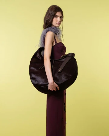 Fashion model in a burgundy dress with lilac feather trim holding an oversized dark crescent bag against a yellow seamless backdrop.