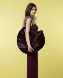 Fashion model in a burgundy dress with lilac feather trim holding an oversized dark crescent bag against a yellow seamless backdrop.