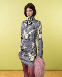 Model wearing a grey floral high-neck mini dress, burgundy tights, and carrying a pink faux-fur accessory against a yellow wall.