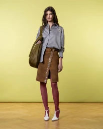 Model wearing a grey button-down shirt and brown skirt carrying an olive green shoulder bag against a gradient yellow background