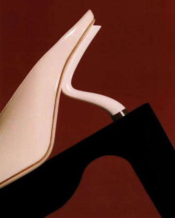 Close-up studio shot of a white heeled mule shoe resting against a black sculptural geometric object on a dark red background.