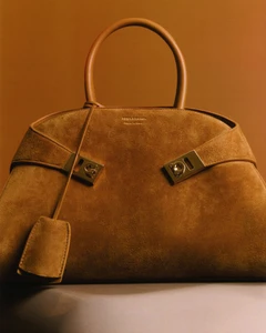 Tan suede handbag with a top handle and metallic hardware centered against a warm brown gradient background.