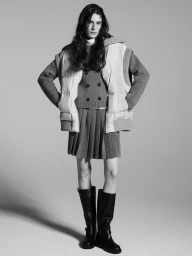 Model wearing a puffer vest, double-breasted knit cardigan, pleated skirt, and knee-high boots against a grey seamless background