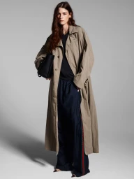Model standing against a grey studio backdrop wearing an oversized trench coat and carrying a black handbag